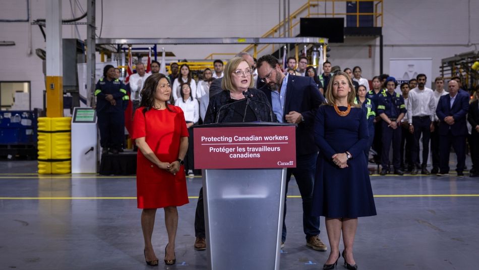 Three people standing at a podium in a factory with a group standing in the background