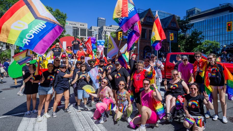 A large group at the Toronto Pride parade waving Pride flags in front of a colourful Unifor float