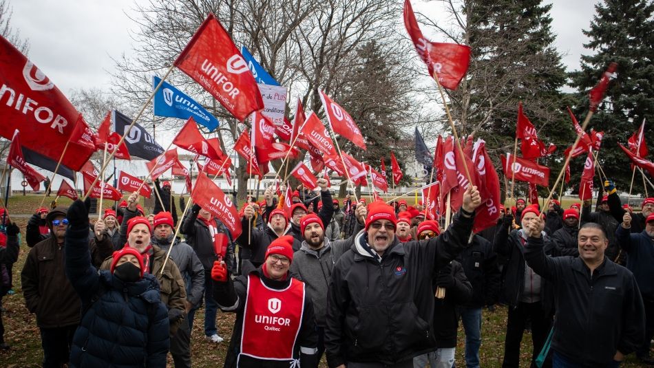 A croud of people ralling and chanting holing up their arms and red Unifor flags.
