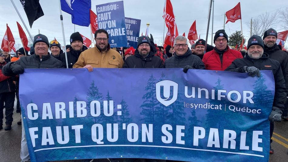 Un groupe de six hommes portant une bannière bleue d'Unifor sur laquelle on peut lire : "Caribou : Faut qu'on se parle". Un groupe de personnes marche derrière eux, agitant des drapeaux rouges d'Unifor.