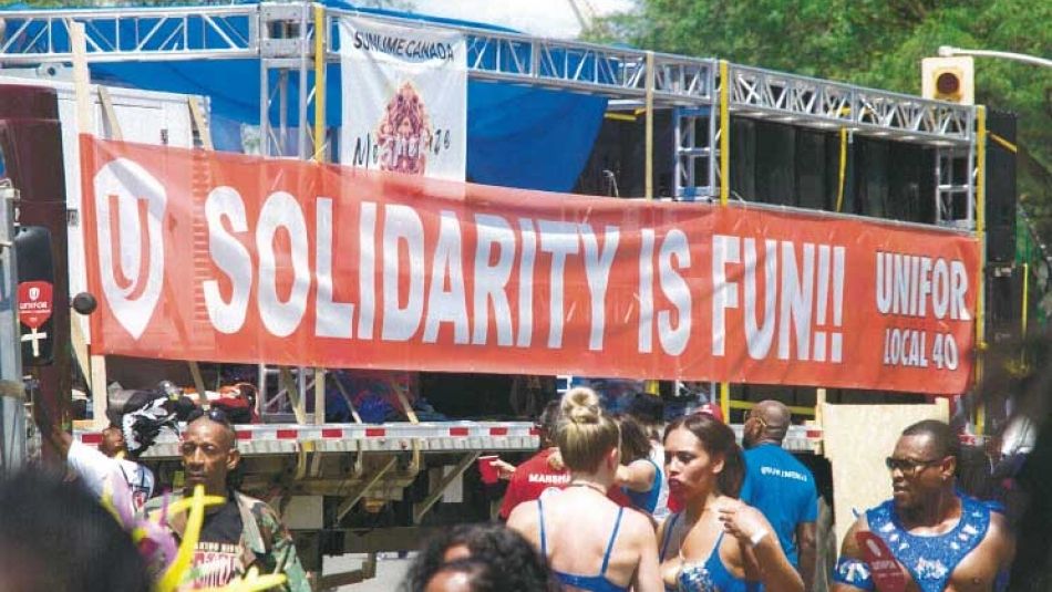 Des participants au Carnaval des Caraïbes de Toronto 2019 avec une bannière rouge d’Unifor en arrière-plan.