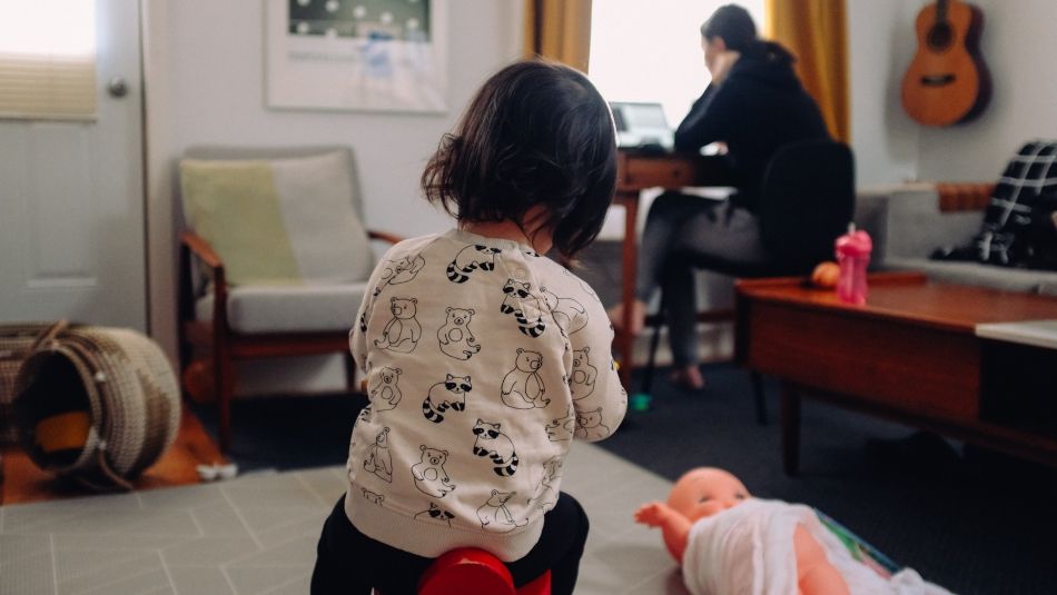 a child sitting on a toy with a parent on a computer in the background