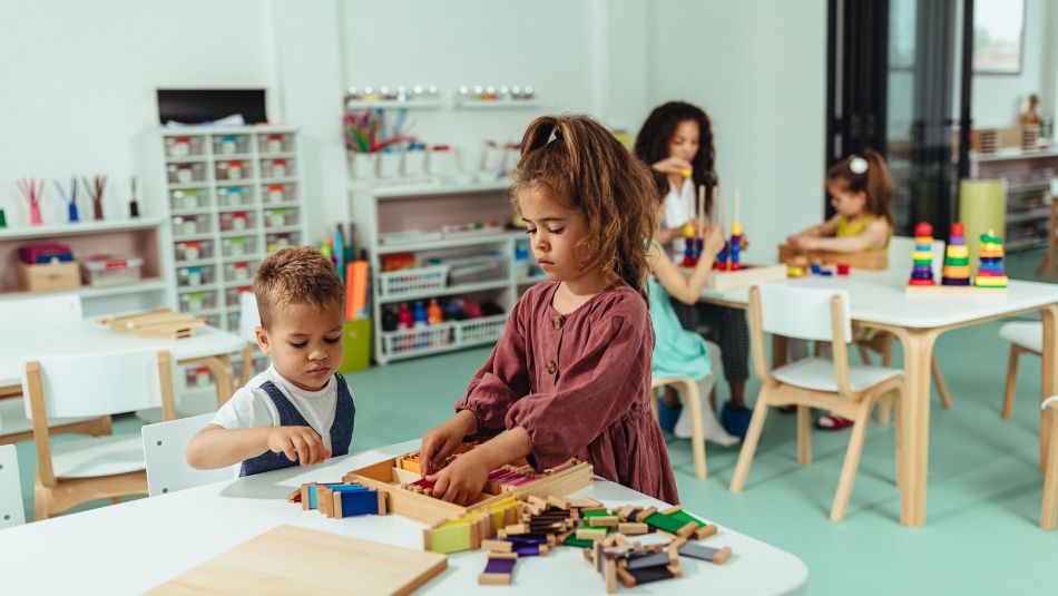 children playing blocks at a table. A women sits with a child in the background