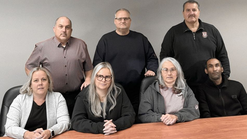 A group photo of the Unifor Local 2458 Clear Medical Imaging bargaining committee, with seven members seated around a wooden table and three standing behind, wearing casual clothing, including some in Unifor-branded attire.