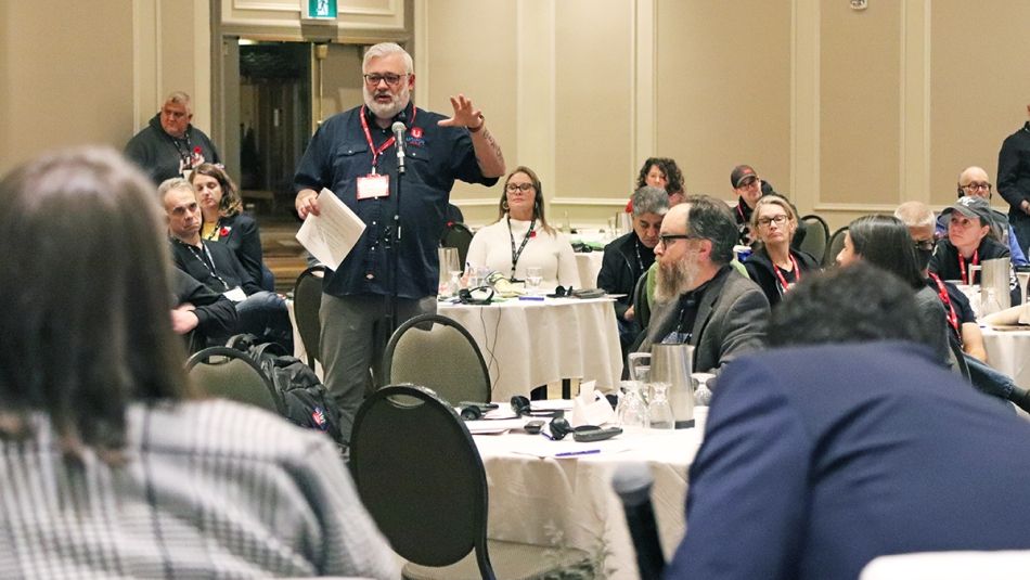 Person standing and speaking at microphone surrounded by people seated at round tables.