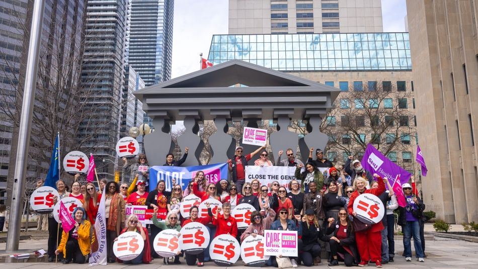 A large group of people pose outside a building, holding signs and banners advocating for closing the gender pay gap and wage equality.