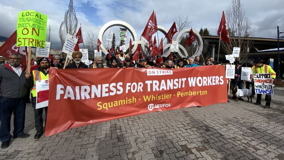 Grande foule devant les anneaux olympiques de Whistler tenant des drapeaux et une grande bannière « Équité pour les travailleurs du transport en commun »