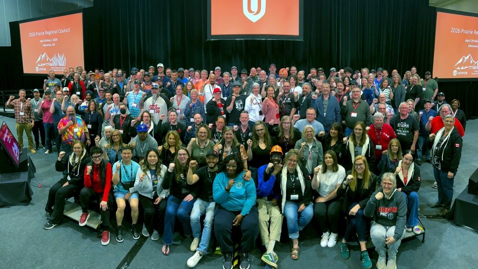 More than one hundred delegates posing in front of a stage with Unifor logo on screens behind.