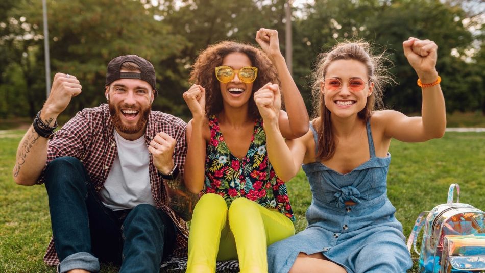 Three smiling friends sit at bright, lush park on grass.