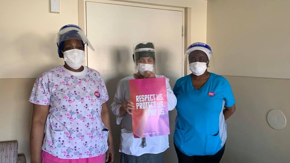 three diverse health care workers wearing masks and faceshields the women in the middle is holding a respect us, protect us, pay us sign