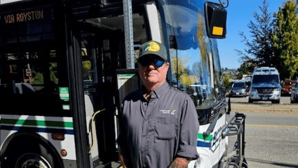 A man in a hat and sunglasses stands infront of a transit bus