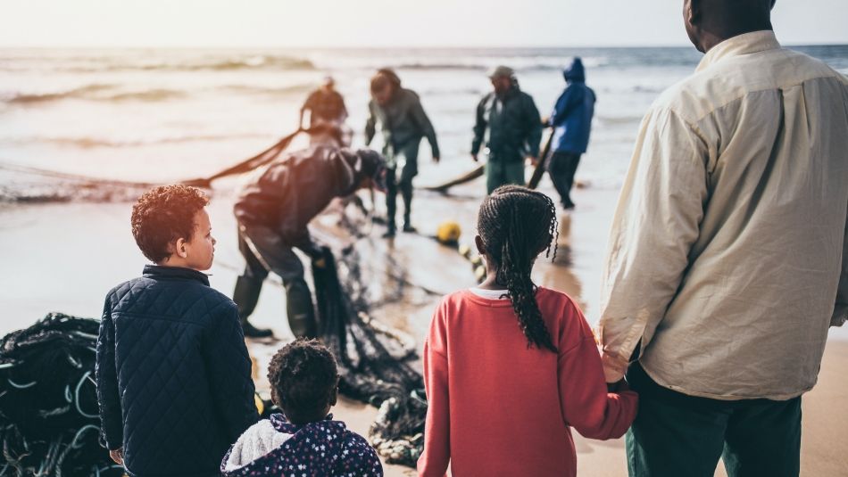 A black father holds the hand of his three children, watching men pull nets out of the sea.