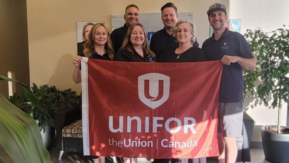 Three women standing and three men standing in a row behind holding up a red Unifor flag