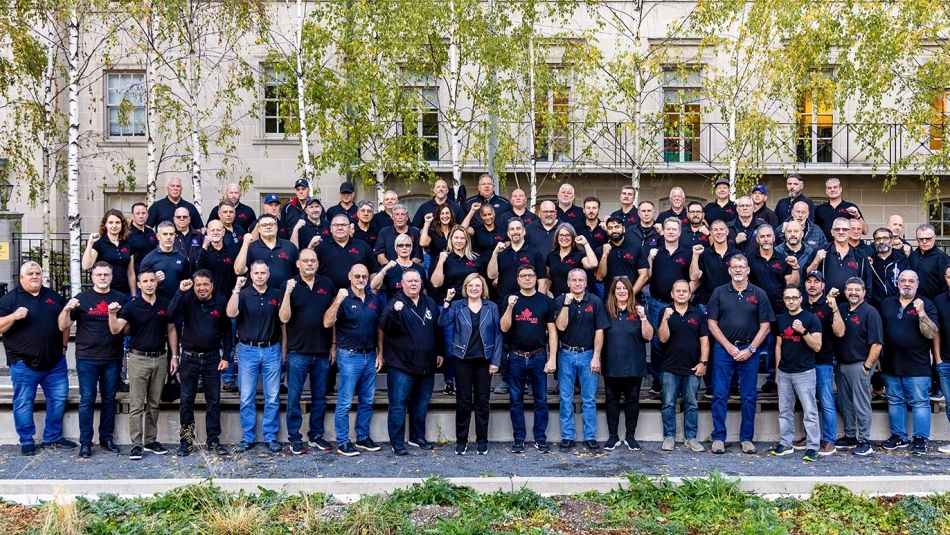 Large group of workers posing for a photo outside in three rows wearing dark t-shirts and holding their fists in the air.
