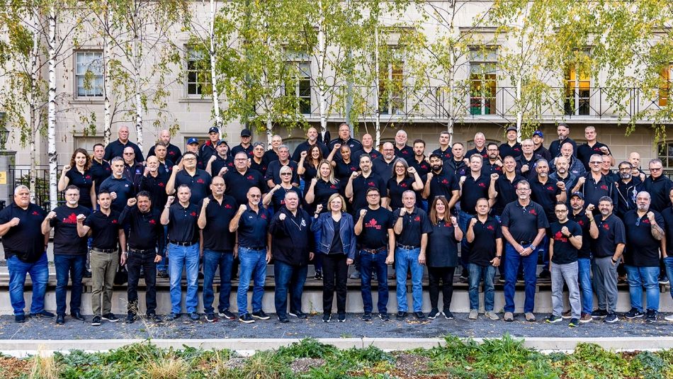 Large group of workers posing for a photo outside in three rows wearing dark t-shirts and holding their fists in the air. 