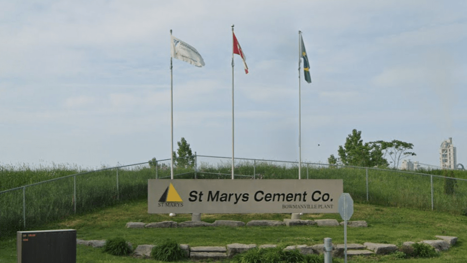hree flags behind a company sign, "St Marys Cement Co." with a chainlink fence and overgrown grass in the background.