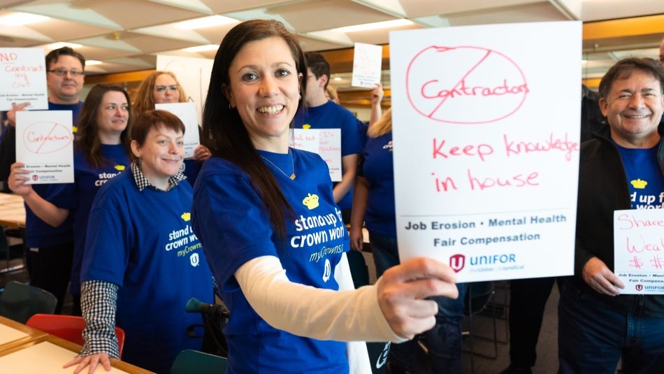 Smiling woman in blue t-shirt holding small sign reading No Contracting Out; Keep Knowledge In House. Other coworkers wearing blue shirts in background. 