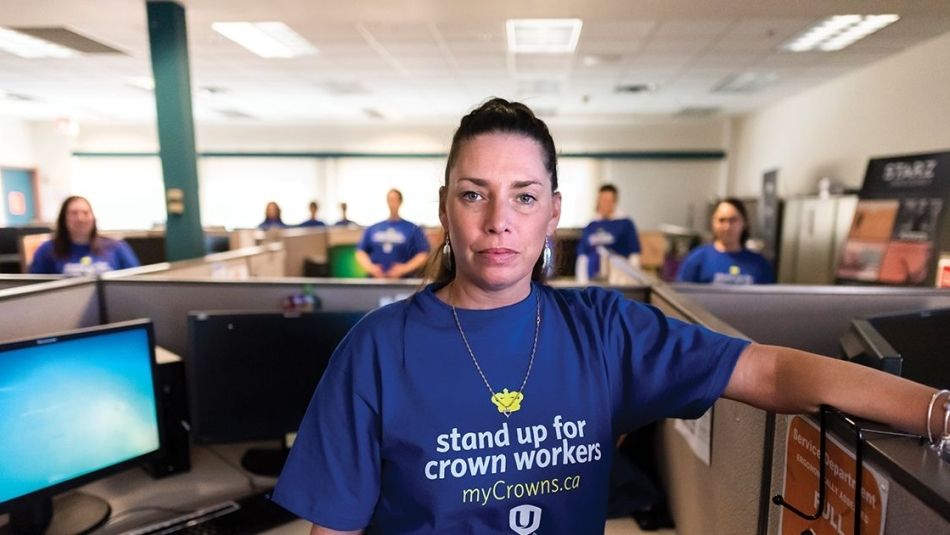 A woman wearing a blue shirt stands in the centre of an office, with people in the background.