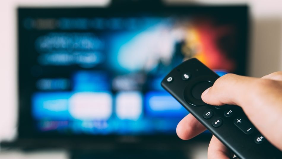 A persons thumb on a TV remote control with a blured out TV in the background.