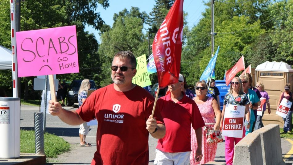 Unifor members walk a picket line carrying flags and signs. One sign reads "Scabs go home."