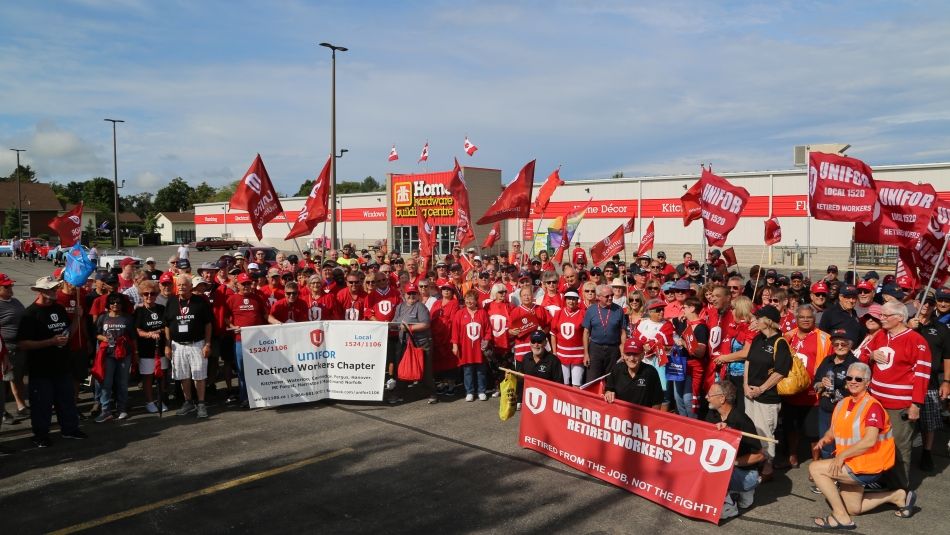A large group of Unifor retirees wearing matching shirts and waving flags in front of a Home Hardware store.