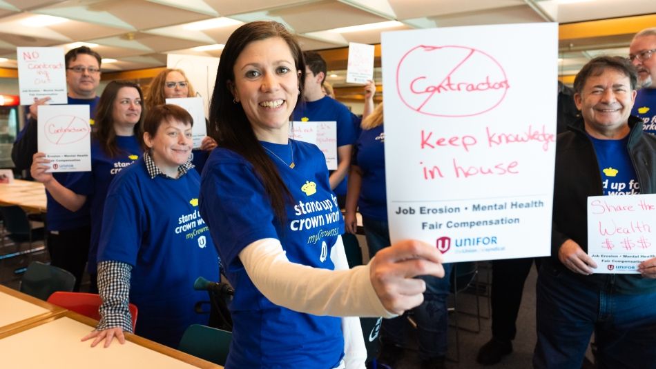 Femme souriante brandissant une pancarte avec le slogan "No Contractors, Keep Knowledge in house" (Non aux sous-traitants, gardez le savoir-faire chez nous). D’autres personnes revêtues du même chandail bleu sont rassemblées autour d’elle.