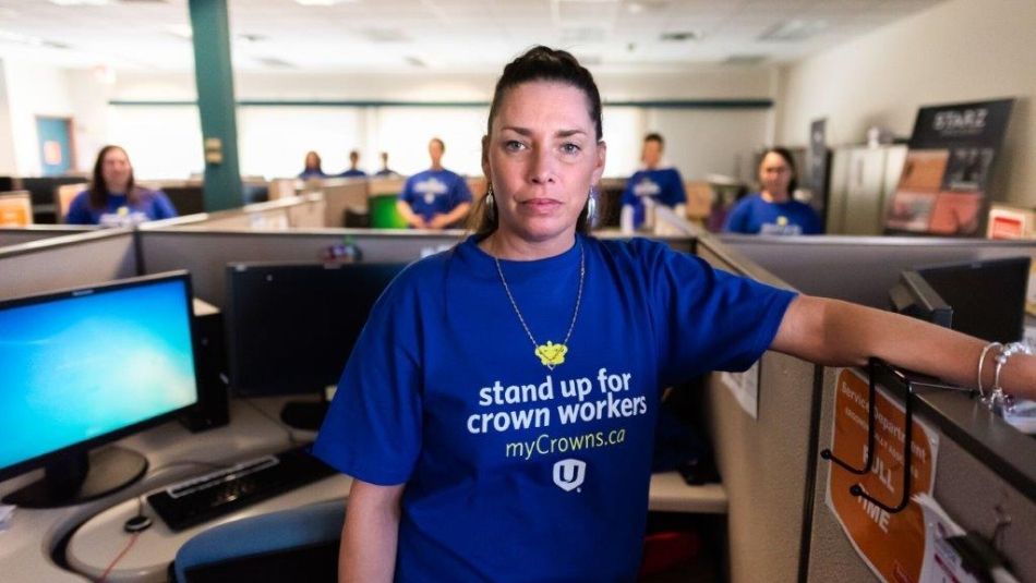 A Unifor member stands in their workspace wearing a blue t-shirt in the background several other members stand in their work spaces wearing identical t-shits.