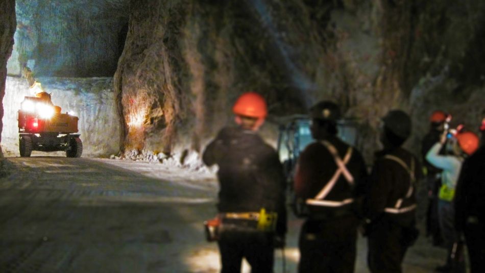 Three miners underground looking down a dark tunnel at a truck operating on the wall of a mine.