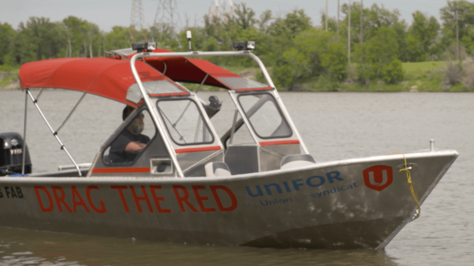 Video screenshot of an aluminum boat with a red canopy on the Red River. The boat says Drag the Red on the side and has a Unifor logo on the hull.