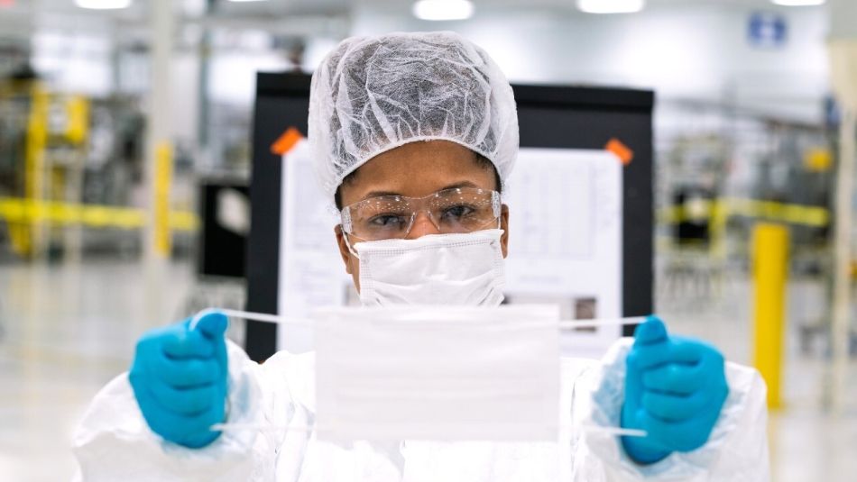 A worker in full PPE holds up a medical mask.