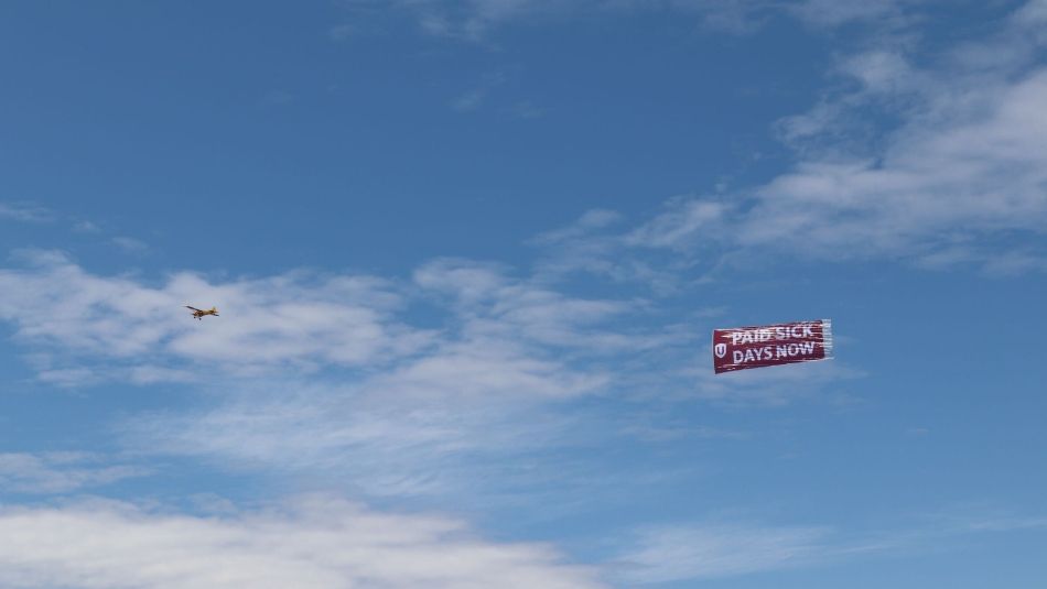 An airplane flys, pulling a banner that reads, "Paid Sick Days Now."