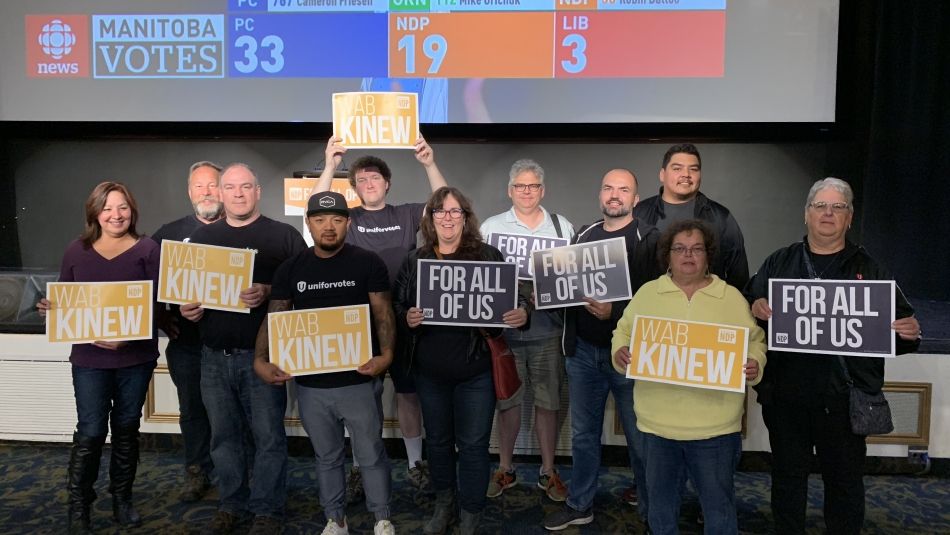 A group of Unifor members hold signs reading "For all of us" and "Wab Kinew."