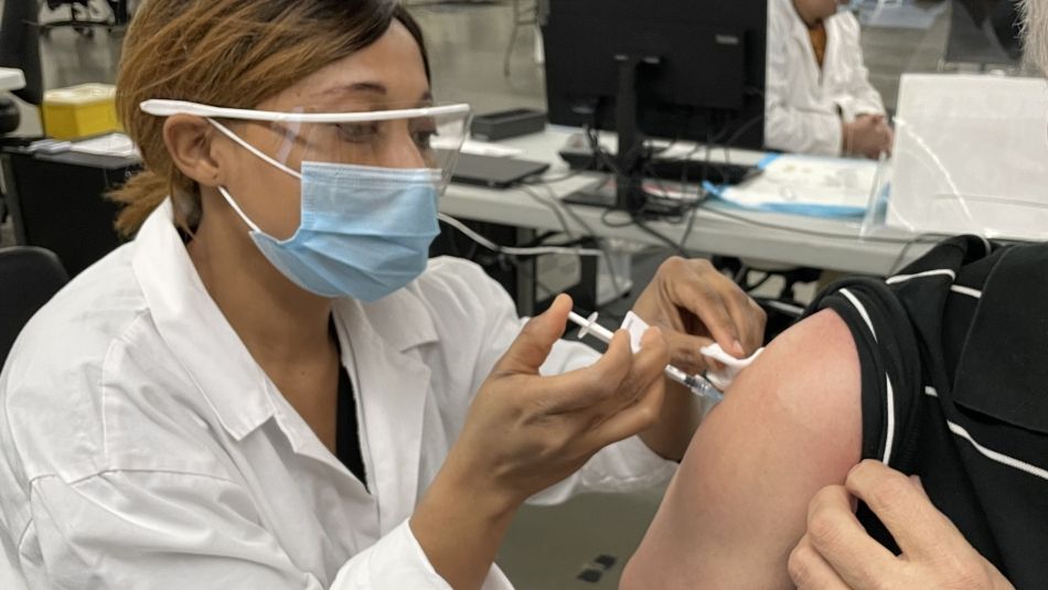 Health professional in goggles and mask giving COVID vaccine needle to seated man.