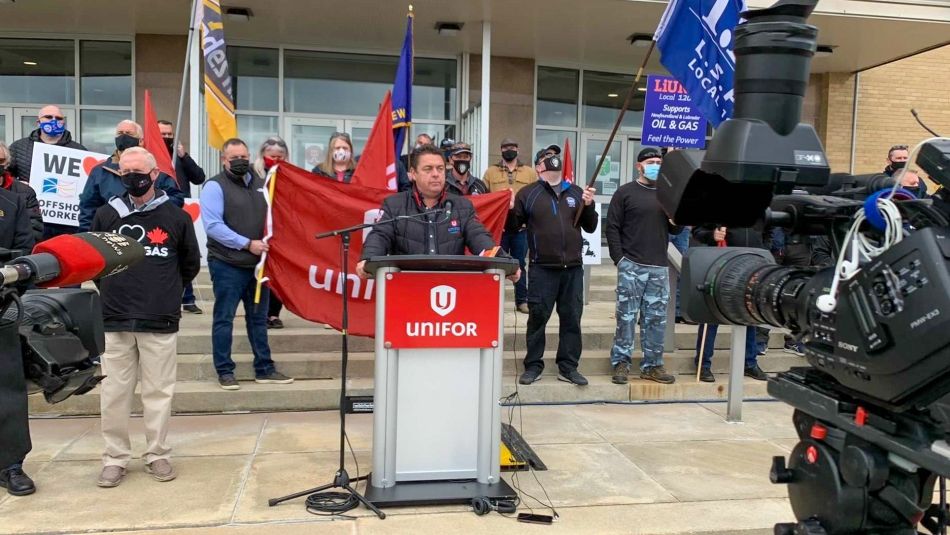Dave Mercer from Unifor Local 2121 standing at podium with demonstrators behind him and TV cameras in the foreground.