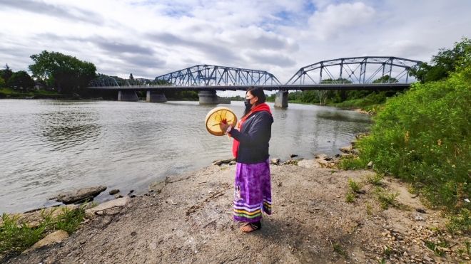 Woman standing on riverbank holding a drum. A bridge is visible in the background.