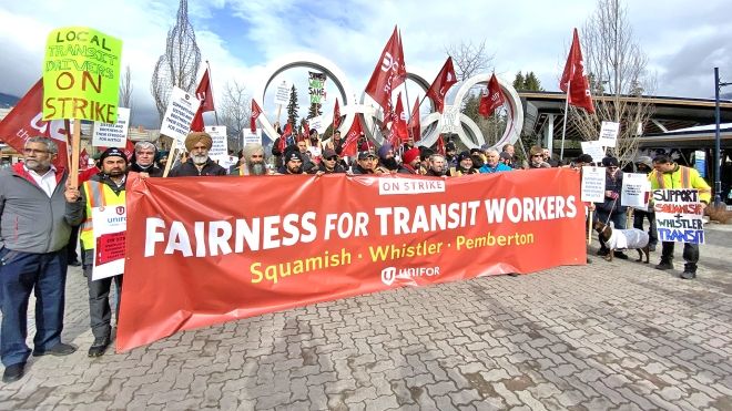 Large group of rally participants with flags and placards posing with a "Fairness for Transit Workers" banner in front of Whistler Village's Olympic rings..