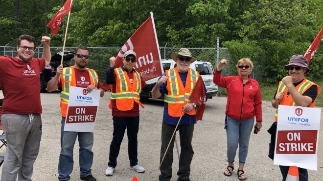 Six Unifor members standing on the road outside an Enbridge facility with hi-viz vests, red shirts, flags, and an On Strike placard.