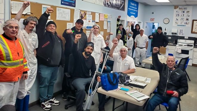 "18 workers posing for a photo with fists in the air in a lunch room. Some wearing white uniforms and hair nets."
