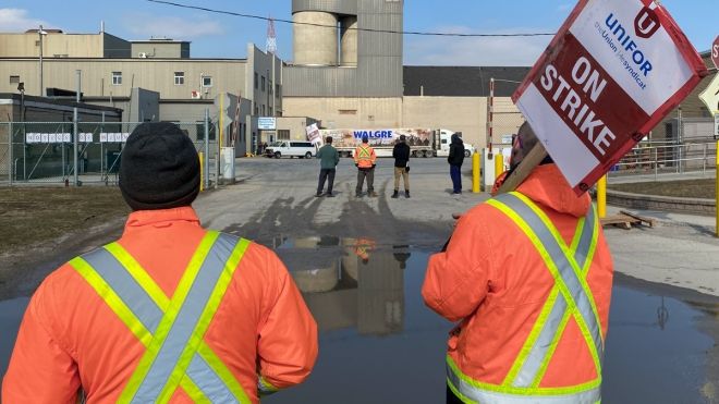 Two workers on picket line in hi viz jackets facing Windsor Salt facility in distance.