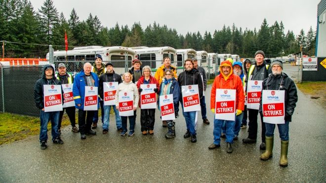 A group of workers wearing "on strike" placards posing for a photo on a driveway with several buses parked in the background. 