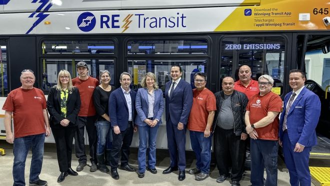A dozen people post for a photo standing beside a bus inside a warehouse.