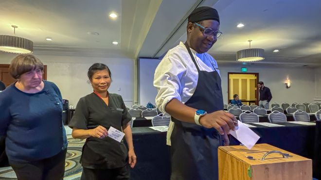 One worker puts paper into a ballot box while two are lined up behind him.