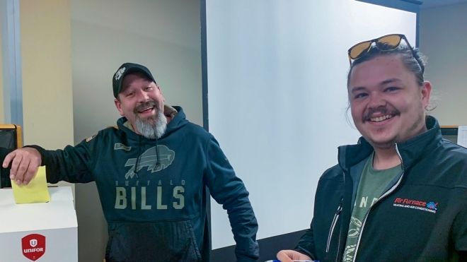 Two people standing and smiling, one is depositing a ballot in a ballot box.