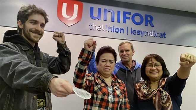Four people indoors with their fists in the air. Unifor signage on the wall behind them