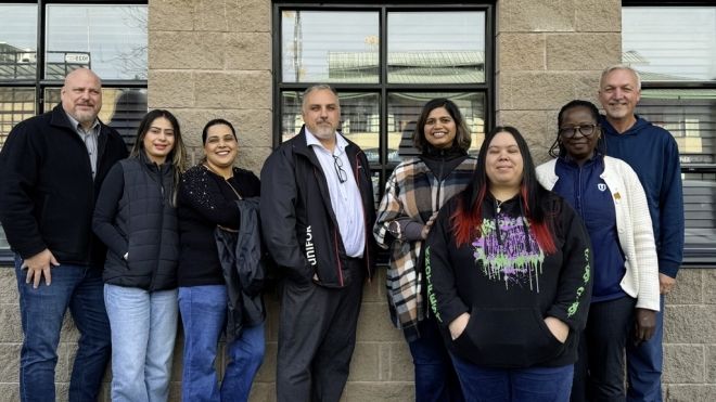 Eight people standing outside near the side of a brown building. 