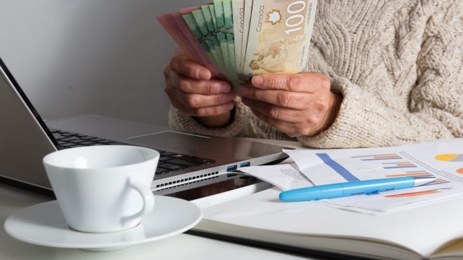 a person sitting at a laptop next to a notebook and a cup of tea holding money different bill fanned out