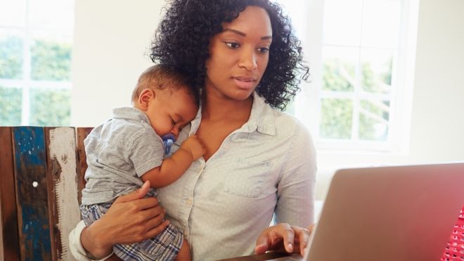 A women holds a young baby while working at a computer.