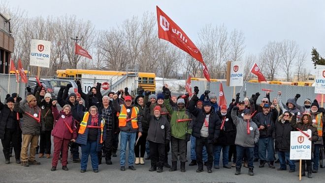 A group of people, raising their firsts in the air and holding white Unifor placards in front of a chain-link fence with yellow school buses in the background.