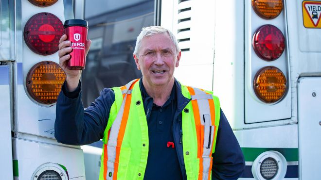 A man in a yellow safety vest holds up a Unifor travel mug in front of a BC transit bus