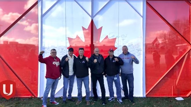 Six men standing in front of a large Canadian flag signage fists raised
